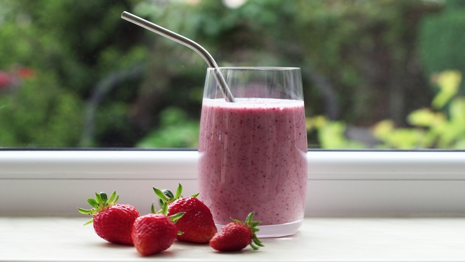 Batido de frutillas en un vaso con sorbete metálico, junto a frutillas frescas en el borde de una ventana